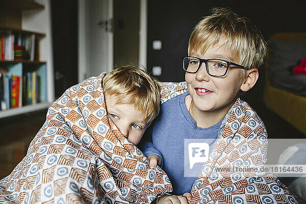 Happy boy with brother wrapped in blanket sitting at home
