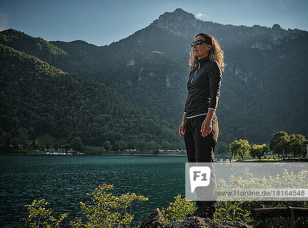 Reife Frau mit Sonnenbrille steht auf einem Felsen am See