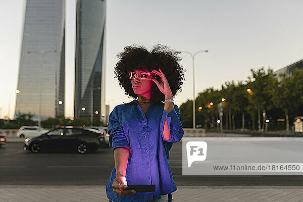 Red light falling on Afro woman face wearing smart glasses holding smart phone on road