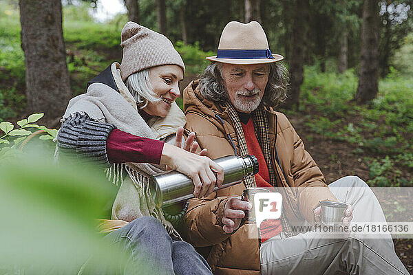 Mature woman pouring water from thermos by senior man in forest