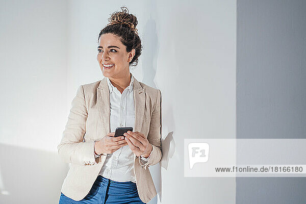 Smiling businesswoman with smart phone leaning on wall