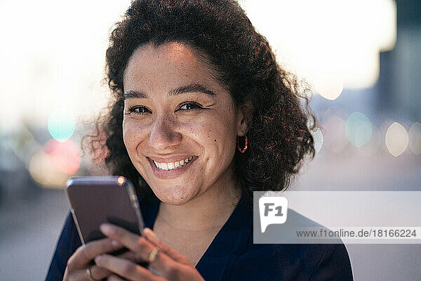 Happy curly hair businesswoman with mobile phone at dusk