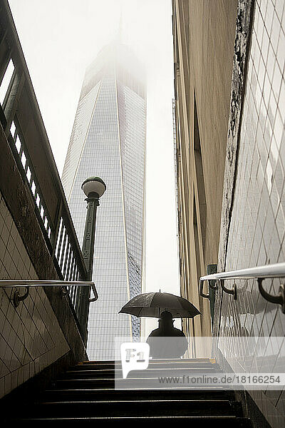 USA  New York  New York City  Pedestrian standing with umbrella in front of subway entrance with skyscraper shrouded in fog in background