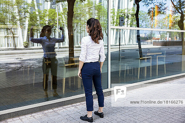 Businesswoman looking at reflection flexing muscles on glass wall