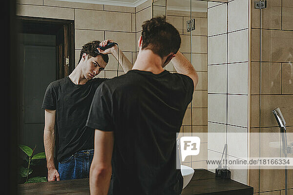 Young man with electric razor cutting his hair in bathroom