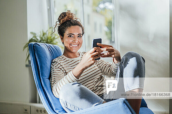 Smiling woman holding mobile phone on chair at home