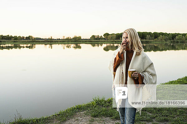 Woman holding cup eating apple by lake at sunset