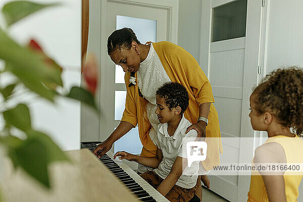 Girl looking at mother teaching son to play piano at home