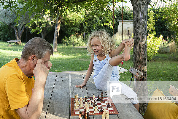 Happy blond girl looking at grandfather playing chess on table in garden