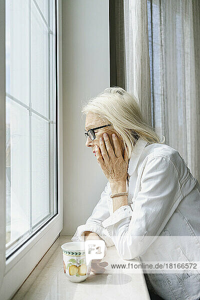Deprimierte ältere Frau mit Tasse lehnt zu Hause auf Fensterbank