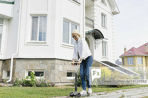 Happy senior woman riding push scooter in front of house