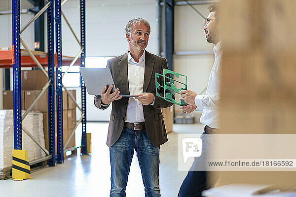 Senior businessman holding laptop talking to colleague standing in warehouse