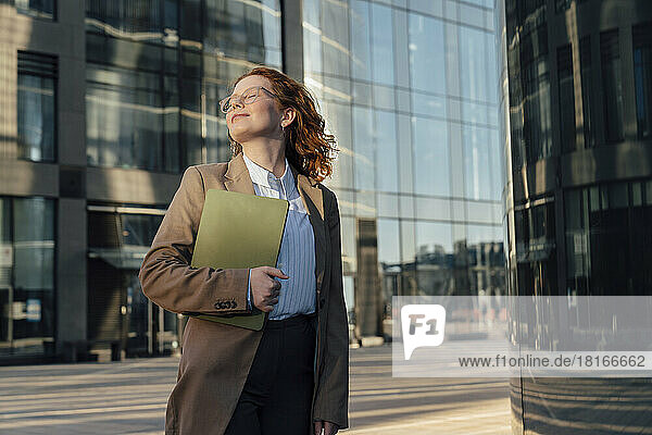 Smiling businesswoman with eyes closed and laptop by office building