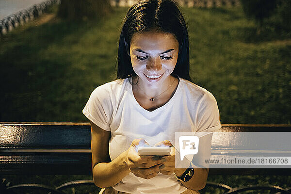 Happy woman using smart phone sitting on park bench at night