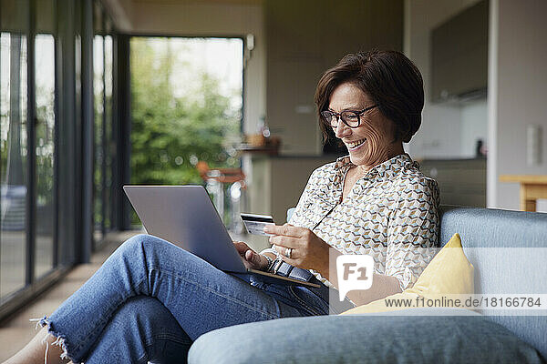 Happy woman paying with credit card through laptop on sofa at home
