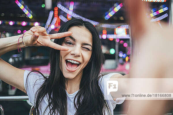 Cheerful woman winking and showing peace gesture at amusement park
