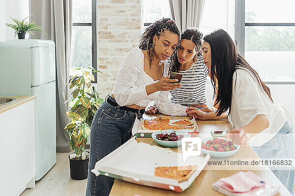 Young woman sharing smart phone with friends in kitchen