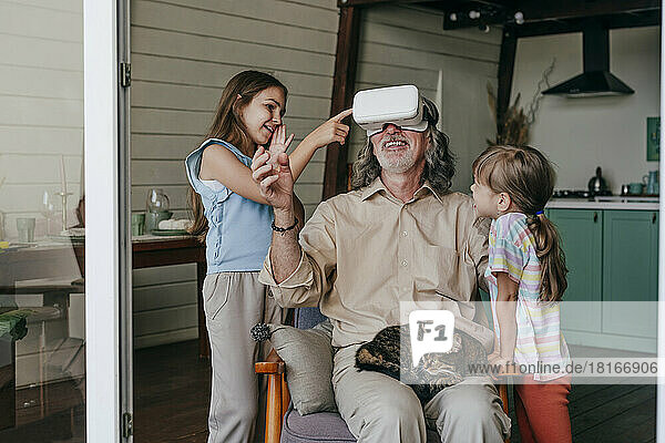 Lächelnder Großvater mit VR-Brille gestikuliert von Enkelinnen zu Hause