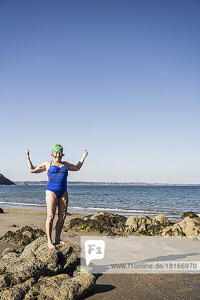 Frau steht auf einem Felsen und lässt am Strand ihre Muskeln spielen