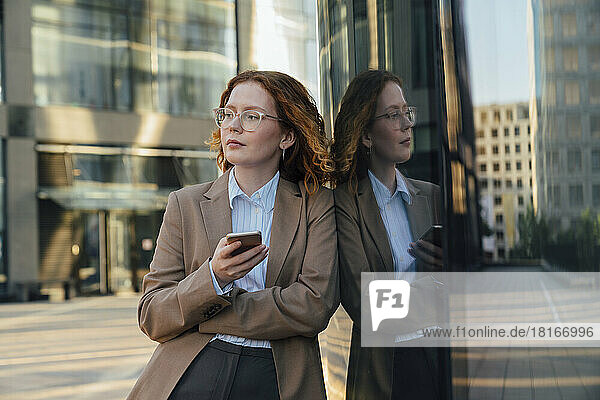 Thoughtful businesswoman with smart phone leaning on glass wall