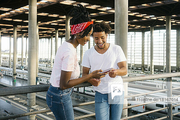 Happy multiracial friends using smart phone together at railroad station