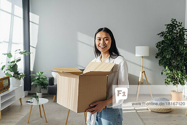 Lächelnde Frau hält einen Karton im Wohnzimmer ihres neuen Zuhauses