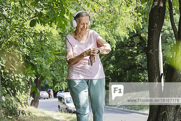 Smiling senior woman wearing wireless headphones walking by tree
