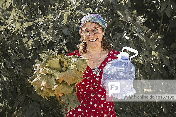 Lächelnde Frau hält Salat und Plastikgallone vor Pflanzen