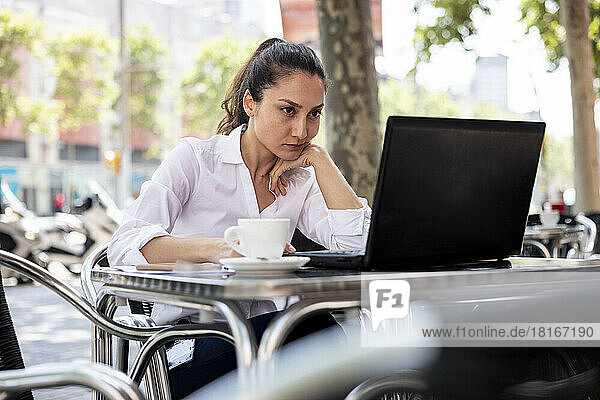 Businesswoman with hand on chin working on laptop at sidewalk cafe