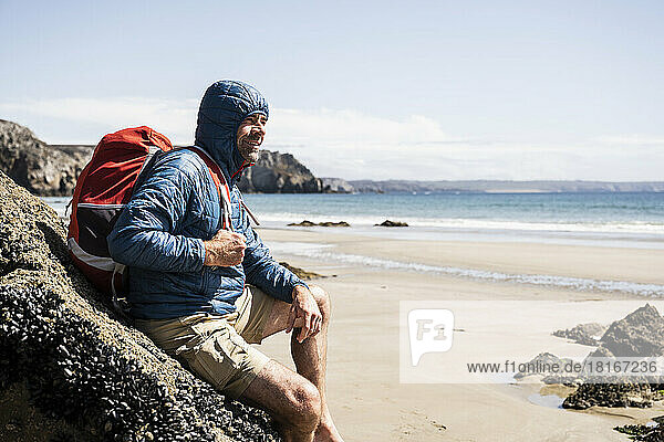 Nachdenklicher reifer Mann mit Rucksack  der am Strand auf einem Felsen lehnt
