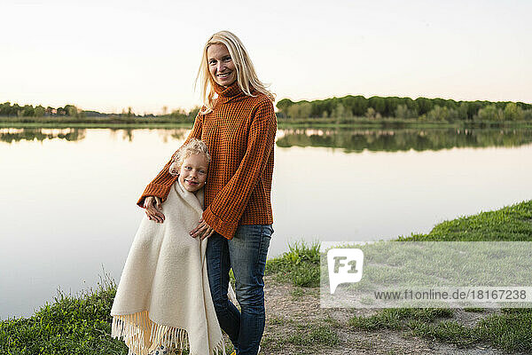 Smiling woman standing with daughter wrapped in blanket at lake