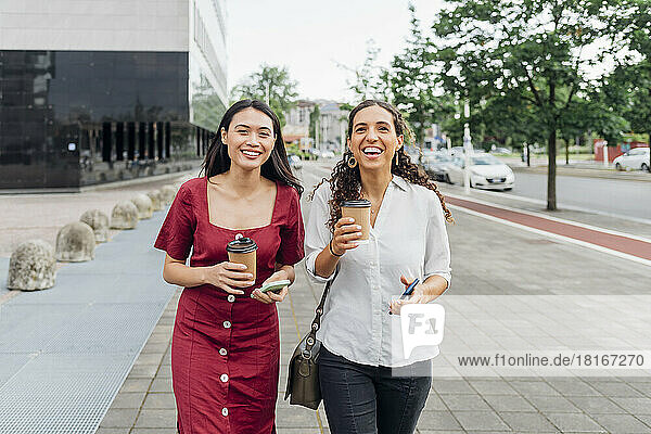 Happy young friends with reusable cups walking on footpath