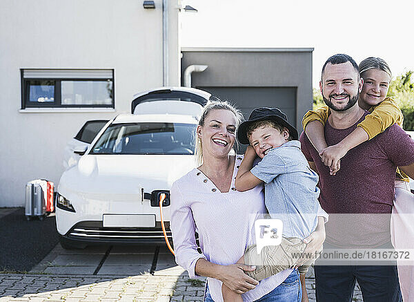 Happy family standing in front of family home and charging electric car