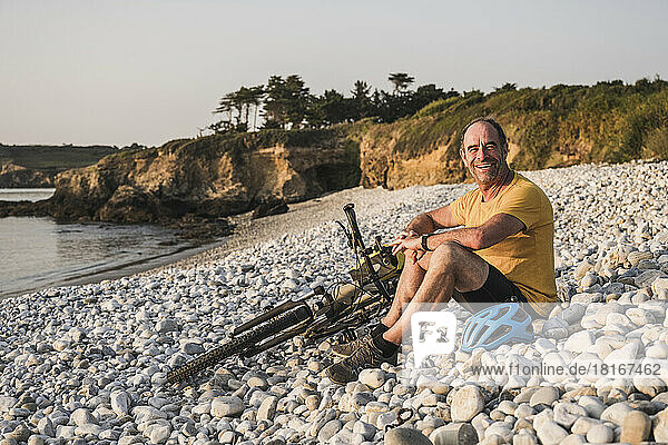 Happy mature man sitting with bicycle at beach