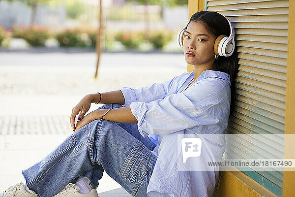 Young woman wearing headphones listening to music leaning on shutter