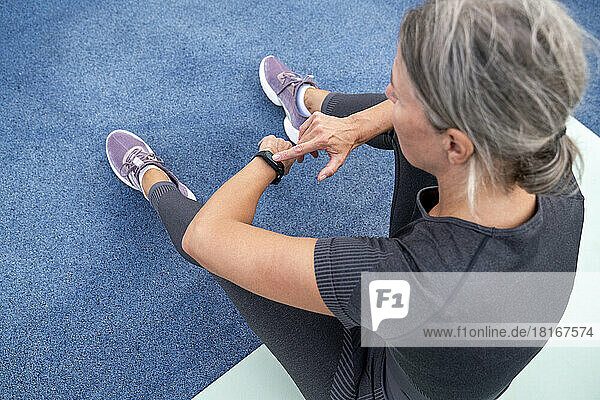Reife Frau überprüft die Uhrzeit mit einer Smartwatch  die auf einer Trainingsmatte sitzt
