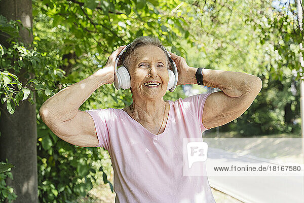 Happy senior woman wearing wireless headphones exercising in front of tree