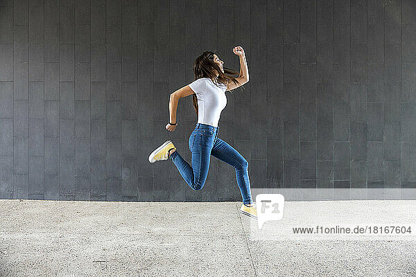 Playful woman jumping on footpath in front of gray wall