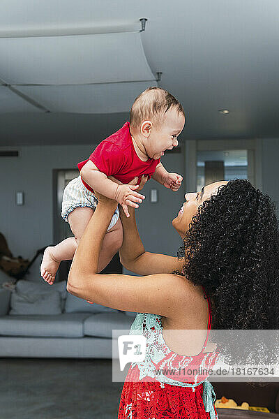 Happy woman with curly hair holding son aloft at home