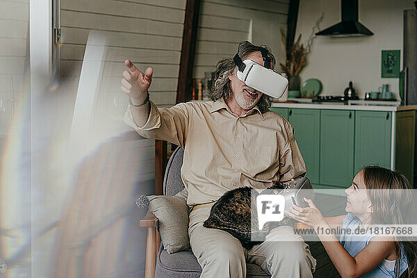 Happy grandfather with VR glasses gesturing with granddaughter at home