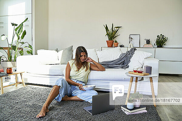 Woman holding bowl looking at laptop by sofa in living room