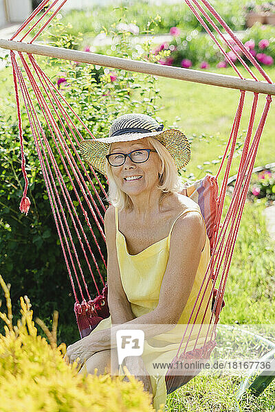 Smiling senior woman wearing hat sitting on hanging chair in garden