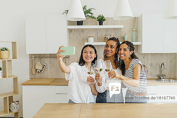 Happy friends talking selfie with wineglasses in kitchen