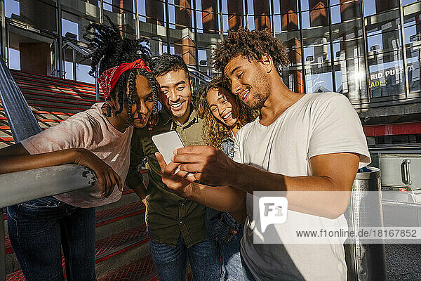 Smiling man sharing mobile phone with multiracial friends at railroad station