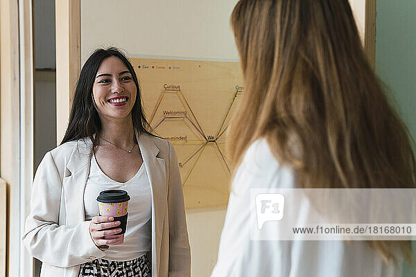 Lächelnde Geschäftsfrau mit Einwegkaffeetasse blickt Kollegin im Büro an