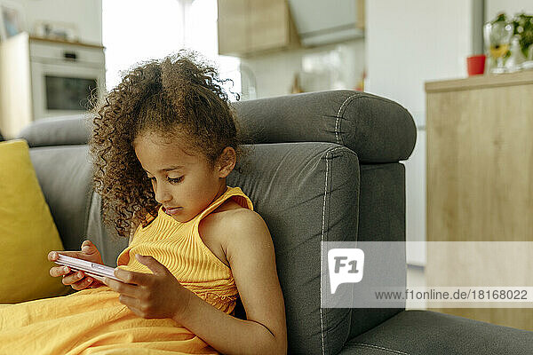 Girl with curly hair watching video through mobile phone on sofa at home