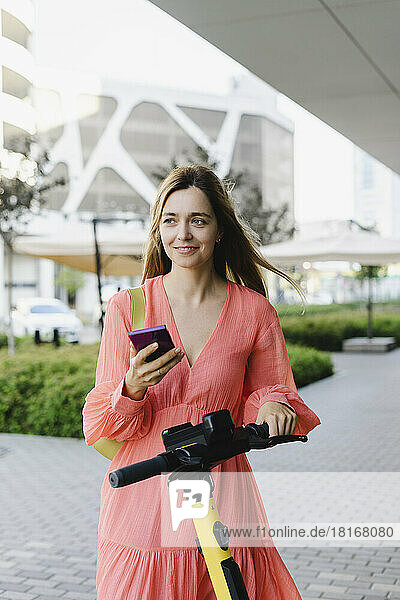 Smiling woman holding smart phone standing with electric push scooter on footpath