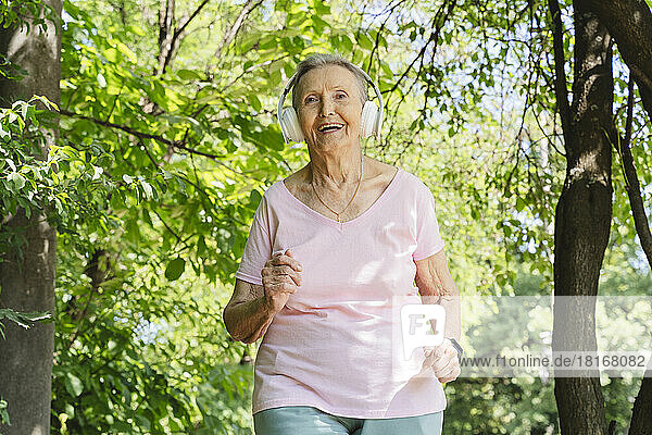 Glückliche aktive Seniorin mit kabellosen Kopfhörern joggt inmitten von Bäumen
