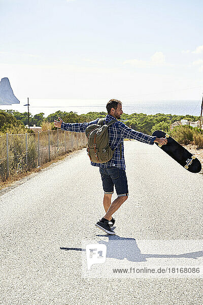 Backpacker with arms outstretched carrying skateboard on sunny day