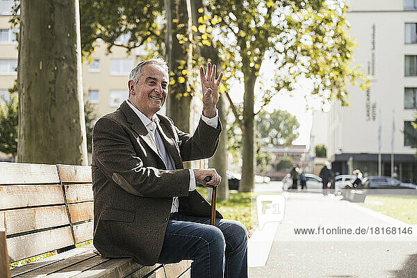 Smiling senior man with walking cane waving and sitting on bench